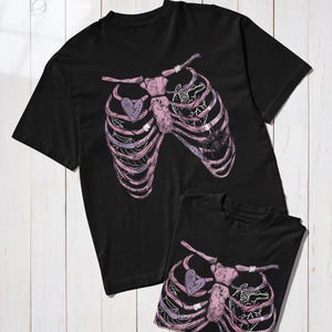 Puede incluir: Dos camisetas negras con un dise&ntilde;o de caja tor&aacute;cica en tonos rosas y morados, con un coraz&oacute;n y un gr&aacute;fico de ara&ntilde;a. Las camisetas se muestran sobre una superficie de madera blanca.