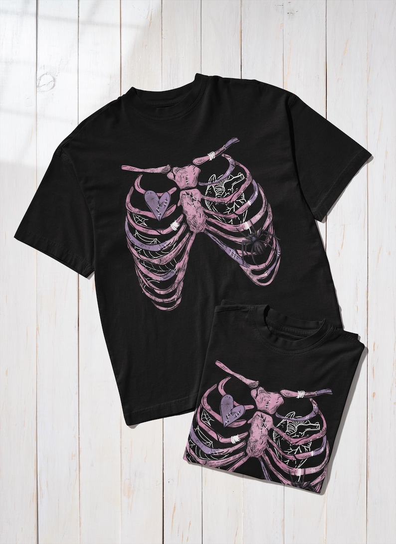 Puede incluir: Dos camisetas negras con un dise&ntilde;o de caja tor&aacute;cica en tonos rosas y morados, con un coraz&oacute;n y un gr&aacute;fico de ara&ntilde;a. Las camisetas se muestran sobre una superficie de madera blanca.