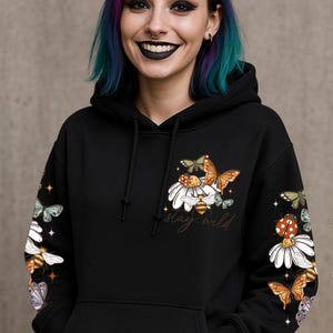 May include: Black hoodie with a floral and insect design. The design includes butterflies, daisies, and bees, with the words "stay wild" in script. The sleeves also feature the same design. The hoodie has a drawstring hood and a front pocket.