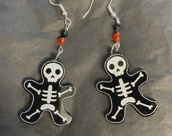 Puede incluir: Un par de pendientes de Halloween. Cada pendiente presenta una figura de esqueleto negro con detalles óseos blancos. Los pendientes tienen ganchos plateados y están acentuados con cuentas rojas y negras.