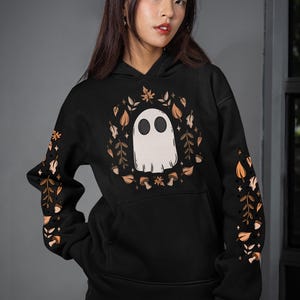 Könnte beinhalten: Schwarzer Kapuzenpullover mit einem weißen Geisterdesign, umgeben von Herbstblättern und Pilzen. Die Ärmel haben passende Blatt- und Pilzmuster. Ein gemütliches, saisonales Kleidungsstück für Halloween oder den Herbst.