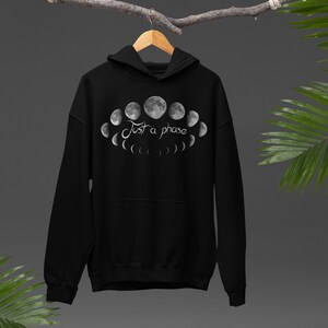 May include: Black hooded sweatshirt with a graphic of the moon phases in grey and white. The text "Just a phase" is above the moon phases. The sweatshirt has a front pocket and a hood.