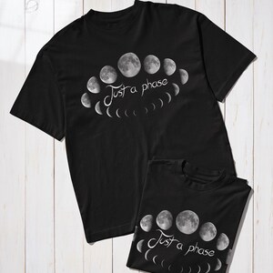 Op de afbeelding: Twee zwarte t-shirts met een witte afbeelding van de maanfasen en de tekst "Just a phase". Het ontwerp toont een reeks maanbeelden in verschillende fasen, waardoor een hemels thema ontstaat.