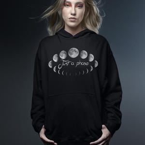 Könnte beinhalten: Schwarzer Kapuzenpullover mit einer weißen Grafik der Mondphasen und dem Text "Just a phase". Das Design zeigt eine Reihe von Mondillustrationen in einem kreisförmigen Muster. Der Pullover hat eine Kapuze mit Kordelzug und eine Kängurutasche.