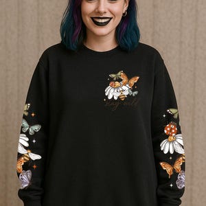 May include: Black sweatshirt with a floral and butterfly design. The design includes daisies, butterflies, and a bee, with the words "stay wild" in script. The sleeves also feature the same design. The sweatshirt is a crew neck.
