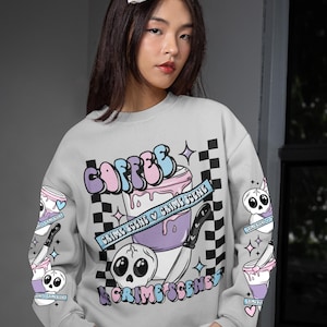 Könnte beinhalten: Ein hellgraues Sweatshirt mit einem grafischen Design. Das Design umfasst das Wort "COFFEE" in Buchstaben, eine Kaffeetasse, einen Totenkopf und die Worte "CRIME SCENE". Die Ärmel haben zusätzliche Grafiken von Kaffeetassen und Totenköpfen.