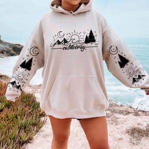 May include: A beige hoodie with a front pocket and a drawstring hood. The hoodie features a black graphic design of mountains, trees, a sun, a moon, and the word "outdoorsy". The sleeves also have similar graphics.