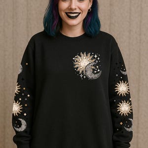Op de afbeelding: Zwarte sweatshirt met een hemels ontwerp. Het ontwerp bevat een gouden zon met een gezicht en een grijze maan, samen met sterren. Het ontwerp staat op de borst en de mouwen. Een casual kledingstuk.