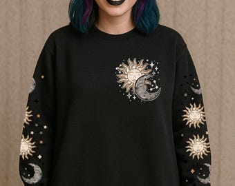 May include: Black sweatshirt featuring a celestial design with a sun and moon graphic on the chest and sleeves. The sun is gold with a face, and the moon is gray. Stars and other celestial elements complete the design.