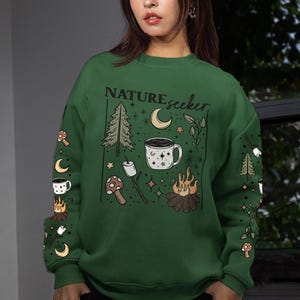 Puede incluir: Sudadera verde con gráficos temáticos de acampada. El diseño incluye una fogata, una taza de café, una luna, árboles y el texto "NATUREseeker". Las mangas presentan ilustraciones de acampada adicionales. Se lleva un pañuelo blanco y azul en la cabeza.