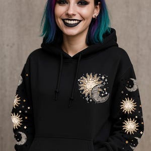 Puede incluir: Sudadera con capucha negra con un diseño celestial. La parte delantera presenta un gráfico de sol y luna con estrellas. Las mangas tienen patrones de sol, luna y estrellas. La persona tiene el pelo colorido y lápiz labial negro.