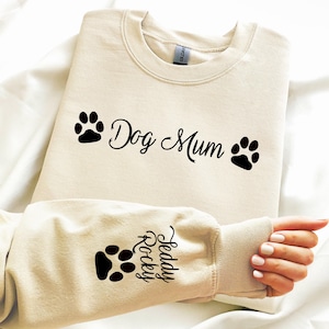 May include: Cream-colored sweatshirt with the text "Dog Mum" and paw prints in black. The sleeve features a paw print and the name "Teddy Rocky". The sweatshirt is made of a soft material.