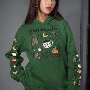 May include: Green hoodie featuring the text "NATUREseeker" and camping illustrations. The design includes a campfire, a mug, a tree, a crescent moon, and a marshmallow on a stick. The sleeves have repeating camping graphics.