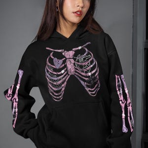 Op de afbeelding: Zwarte hoodie met een roze en paars skelet ribbenkast ontwerp op de voorkant en armen. Het ontwerp bevat een hart en een spin. De hoodie heeft een capuchon en een voorzak.