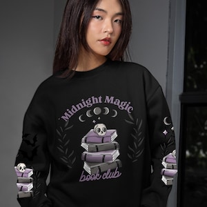 May include: Black sweatshirt with the text "Midnight Magic book club" in purple. The design features a stack of books with a skull on top, crescent moons, and stars. The sleeves have additional book and skull graphics.