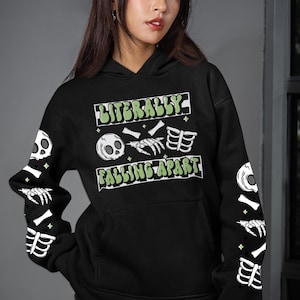 Puede incluir: Sudadera con capucha negra con el texto "LITERALLY FALLING APART" en verde y blanco. La sudadera con capucha presenta gráficos de esqueleto en las mangas y un gráfico de calavera. Tiene un bolsillo delantero y una capucha.