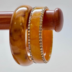 May include: Two round bangles with a smooth, amber-colored marbled finish. One bangle features a solid, polished surface, while the other is accented by two parallel rows of small, clear, sparkling rhinestones embedded along the edges.