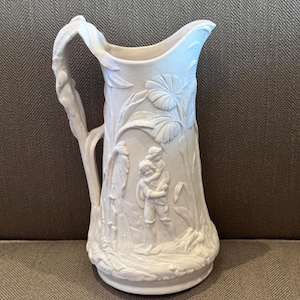 May include: A white ceramic pitcher featuring an embossed relief design of two figures, one carrying the other, surrounded by tropical foliage. The handle is shaped like a leafy vine, and the surface has a matte finish with intricate detailing.