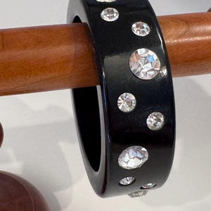 May include: A wide, smooth black bangle bracelet featuring a scattered arrangement of clear, round, faceted rhinestones embedded across the outer surface. The bracelet has a glossy finish and a uniform, thick band construction.