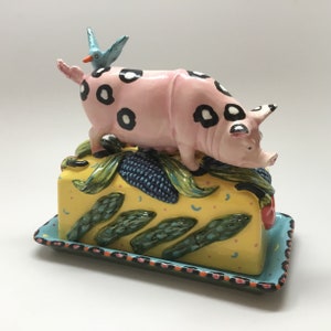 May include: A pink pig with black spots and a blue bird on its back is perched on a yellow butter dish with green and blue floral designs. The dish has a blue and black border.