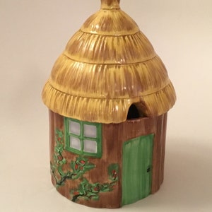 May include: A ceramic cottage-shaped sugar bowl with a thatched roof, green door, and window. The bowl has a brown base with green vines and red berries.