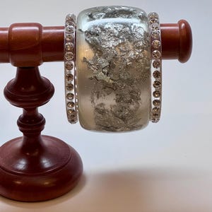 May include: A wide, clear resin bangle bracelet featuring embedded silver-colored metallic flakes, flanked by two thinner, clear resin bangles that are each set with a continuous row of small, round, clear faceted crystals.