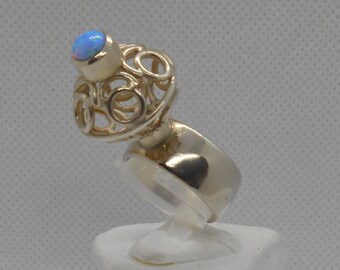 May include: A silver ring featuring a blue opal gemstone set in a delicate, intricate design. The ring is displayed on a white stand.