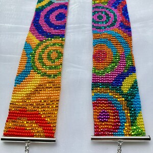 Könnte beinhalten: Zwei bunte Perlenarmbänder mit einem silbernen Verschluss. Die Armbänder zeigen ein geometrisches Muster aus Kreisen in Blautönen, Grün, Gelb, Orange, Rosa und Rot.