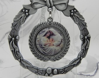 May include: A circular pewter-coloured metal ornament featuring a wreath design with holly leaves and a bow at the top. A central circular medallion depicts a religious scene of a figure holding a baby, surrounded by a textured laurel leaf border.