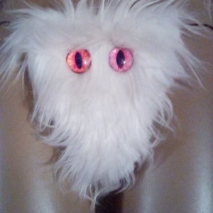 May include: White fluffy toy with two large pink and red eyes. The toy is shaped like a heart and has a furry texture.