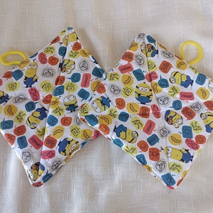 Può includere: Un paio di presine bianche con un bordo giallo. Le presine sono decorate con un motivo di Minion gialli e fumetti colorati. I fumetti contengono frasi come "Banana!" e "Hello!"