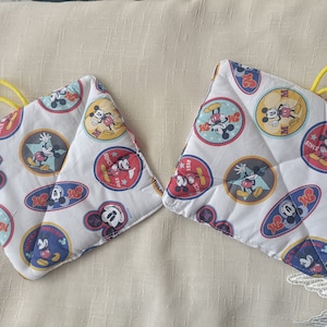 Può includere: Due presine imbottite bianche con tema Mickey Mouse. Le presine presentano varie immagini di Topolino con il testo "Since 1928" e "Mickey Mouse" su di esse.