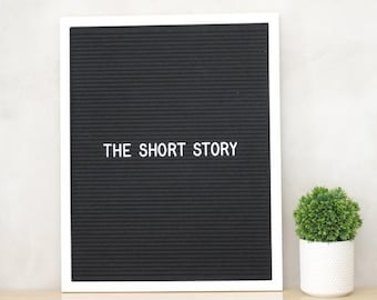Könnte beinhalten: Ein schwarzes Buchstabenbrett mit weißem Rahmen, das auf einer hellen Holzoberfläche steht. Das Brett hat weiße Buchstaben, die "THE SHORT STORY" buchstabieren. Rechts vom Brett steht eine kleine Topfpflanze.