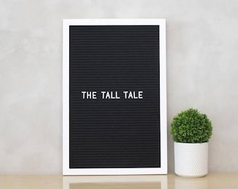 May include: A white framed letter board with black felt letters spelling "THE TALL TALE". A small potted green plant sits on a wooden shelf below the letter board.