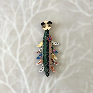 May include: A whimsical, colourful, and handmade wooden figurine of a creature with a face and a long, segmented body. The body is decorated with patterned fabric and a small red flower.