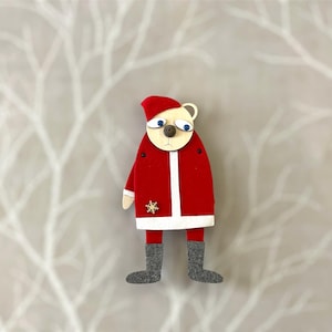 May include: A felt Christmas ornament shaped like a bear wearing a red Santa suit with a white trim and black boots. The bear has a white face with brown eyes and a brown nose.
