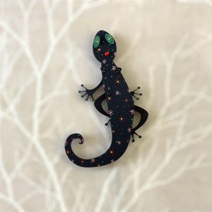 May include: A black gecko with a floral pattern and red and green accents. The gecko is made of wood and has a 3D effect.
