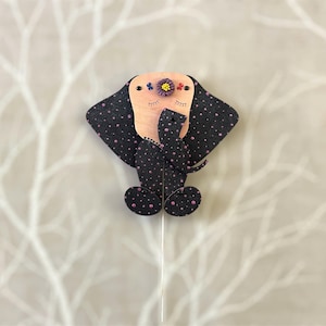 May include: A black and pink polka dot puppet of a dog with a pink nose and closed eyes. The dog has a flower on its head with pink, blue, and yellow petals.