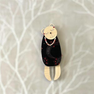 May include: A wooden bear figurine wearing a black and red patterned dress and a pink and white beaded necklace. The bear has a closed-eye smile and is hanging from a string.