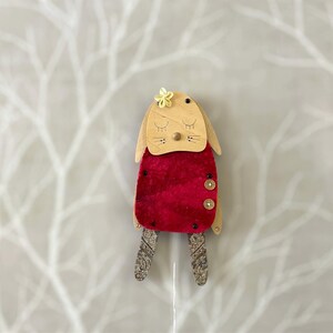 May include: A wooden puppet of a bunny wearing a red velvet dress with a yellow flower on its head. The puppet has a painted face with closed eyes and a small nose.