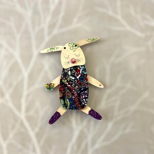 May include: A wooden bunny doll with a colourful patterned dress, green leaves on its ears, and purple shoes. The bunny has a pink nose and closed eyes.