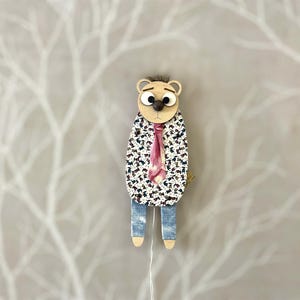 May include: A wooden puppet of a bear wearing a white shirt with a floral pattern and a pink tie. The bear has brown eyes and a brown nose. The puppet is standing on a white background.