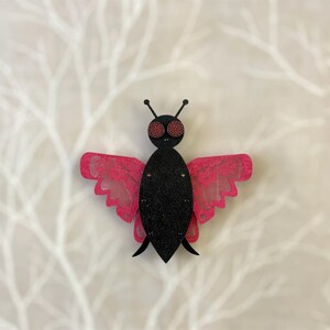 May include: A decorative butterfly ornament. The butterfly is black with glitter and has pink lace wings. The eyes are red with a polka dot pattern. The antennae are black. The item is designed to resemble a butterfly.