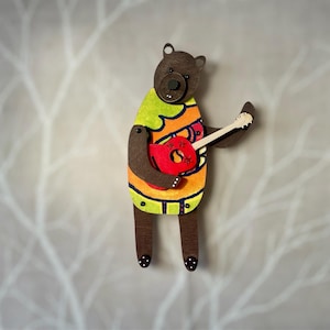 May include: Wooden bear figurine playing a red guitar. The bear wears a colorful shirt with green, orange, and yellow accents. The bear's body is brown, and it has black polka dots on its feet.