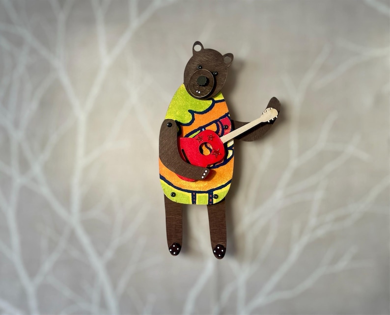 May include: Wooden bear figurine playing a red guitar. The bear wears a colourful shirt with green, orange, and yellow accents. The bear's body is brown, and it has black polka dots on its feet.