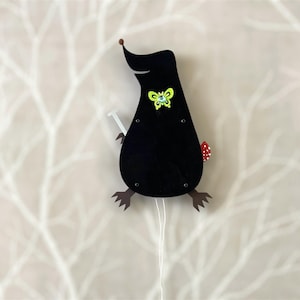 May include: A black wooden toy mole with a green butterfly on its chest and a red mushroom on its tail. The toy is attached to a white string.