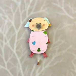 May include: A pink wooden pig with blue and green hearts, light blue wings, and a brown nose. The pig is hanging from a string.