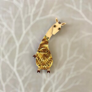 May include: Wooden giraffe ornament with a long neck and a body featuring a crocheted pattern in yellow, brown, and beige. The giraffe has small, dark eyes and ears, and is standing upright.