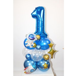Puede incluir: Un globo azul con el número uno, un globo de astronauta, estrellas doradas y globos azules y transparentes. Los globos están dispuestos en forma de torre.