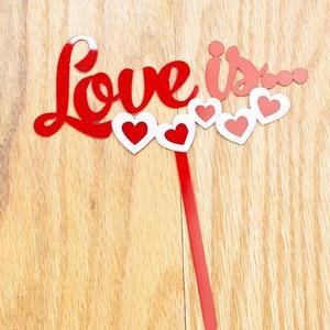 Könnte beinhalten: Roter Acryl-Kuchentopper mit den Worten "Love is..." und einer Reihe roter Herzen.